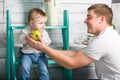 Father gives to baby boy big green apple. He both are in jeans and white hoodie. Dad with son sits on the steps indoor. White