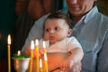 The father hands hold the baby in his arms in the church. The kid looks at the candles burning in the temple.