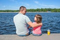 The father having embraced the daughter sit on the embankment against the background of the river in sunny summer day. Family day Royalty Free Stock Photo