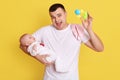 Father holding beanbag toy and animating baby in his hand, posing isolated over yellow background, happy yelling handsome dad Royalty Free Stock Photo