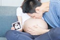 Father is kissing his mother`s stomach. Young husband kissing his pregnant wife`s tummy in living room with the baby ultrasound
