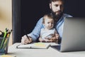 Father with little baby on knees making notes in notebook, looking at laptop. Young man holding child and working on PC Royalty Free Stock Photo