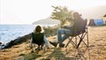 Father with little daughter sits on chairs at camping on lake shore, rear view