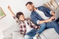 Father and little son at home sitting on sofa dad working on laptop looking surprised at boy holding controller hand up Royalty Free Stock Photo