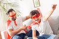 Father and son in superheroe costumes at home sitting on sofa boy close-up hand up shouting motivated while dad holding Royalty Free Stock Photo