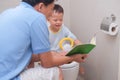 Father reading book to teach smiling son to use toilet in bathroom, Asian 2 -3 years old toddler boy sitting on toilet with kid