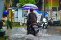 a father riding a motorbike through floodwaters during heavy rain while holding an umbrella in a housing complex