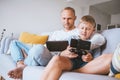 Father and son game players siting together at home on cozy sofa, using the tablet and gamepad on Coronavirus COVID-19 pandemic Royalty Free Stock Photo