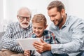 Father, son and grandfather sitting together on couch in living room and looking at digital