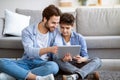 Father and son using tablet computer at home, checking new application, sitting on floor carpet near sofa Royalty Free Stock Photo