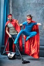 Father in superhero costume vacuuming carpet while son in superhero costume jumping