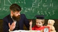 Father teaches son mathematics. Teacher in formal wear and pupil in mortarboard in classroom, chalkboard on background Royalty Free Stock Photo