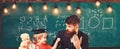 Father teaches son mathematics. Teacher in formal wear and pupil in mortarboard in classroom, chalkboard on background Royalty Free Stock Photo