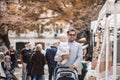 Father walking carrying his infant baby boy child and pushing stroller in crowd of people visiting Sunday flea market in