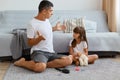 Father wearing white t shirt and jeans shortsitting on floor near sofaand talking with his adorable sad daughter, man having