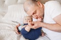 Father in white t-shirt lying in bed with newborn baby son kissing biting his feet toes, Royalty Free Stock Photo