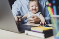 Father working on laptop with baby sitting on his knees.Toddler in front of laptop. Homebased online work for people with children