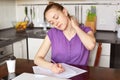 Fatigue adorable woman has pain in neck, being overworked, writes in papers, sits against kitchen inerior with takeaway coffee. Bu Royalty Free Stock Photo