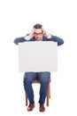 Fatigue businessman seated on chair holding a big blank banner with copy space for messages, keeps hands to head gesture. Full