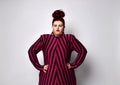 Fatty ginger girl in striped dress and earrings. She has put her hands on waist, pout her lips, posing on white photo background