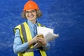 Feamle Engineer in the construction helmet and yellow vest with mobile phone and documents on blue background