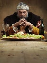 He feasts while the serfs starve. A mature king feasting alone in a banquet hall.