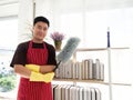 Feather duster in his hand while wearing yellow rubber gloves and sanitizer bottle spray for cleaning book shelf in living room at