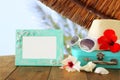 Fedora hat, sunglasses, tropical hibiscus flower next to blank frame over wooden table and beach landscape background