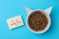 Feeding bowl of kibble with cat ears and cute note For My Lovely Kitty on light blue background, flat lay