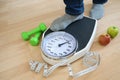 Feet of a man in gray socks stepping on an analog weight scale, green dumbbells, measuring tape and apples lying nearby on a Royalty Free Stock Photo