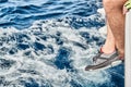 Feet of the sailor dressed in yacht footwear hang down from a boat board over azure water, splashes, sun patches of