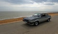 Classic Grey and Silver Ford Capri being driven along seafront promenade beach and sea in background.