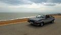 Classic Grey and Silver Ford Capri being driven along seafront promenade beach and sea in background.