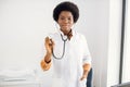 Female African American doctor or nurse, wearing white coat, smiling and demonstrating stethoscope to camera, posing Royalty Free Stock Photo