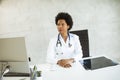 Female African American doctor wearing white coat with stethoscope sitting behind desk in office Royalty Free Stock Photo