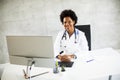 Female African American doctor wearing white coat with stethoscope sitting behind desk in office Royalty Free Stock Photo