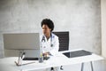 Female African American doctor wearing white coat with stethoscope sitting behind desk in office Royalty Free Stock Photo