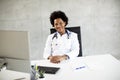 Female African American doctor wearing white coat with stethoscope sitting behind desk in office Royalty Free Stock Photo