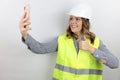 Female architect wearing hardhat smiling on isolated white background holds arm raised holding smartphone and doing thumb up sign. Royalty Free Stock Photo