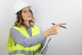 Female architect wearing work helmet and reflecting jacket, holding flip board and pointing with finger to side.