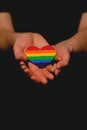 Female arm holding the heart coloured in LGBT pride colours on the dark background. Concept of the International Day Against