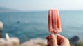 Female arm holding iced strawberry popsicle, blurred sea resort on background, with copy space.