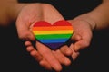 Female arms holding the heart coloured in LGBT pride colours on the dark background. Concept of the International Day Against
