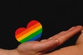 Female arms holding the heart coloured in LGBT pride colours on the dark background. Concept of the International Day Against