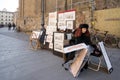 A female artist sells her paintings near the wall of a building in a street lane