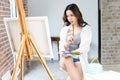 Female Asian artist in casual clothes holding palette and putting brush into paint in studio.Woman painter painting in her Royalty Free Stock Photo