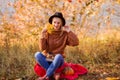 Female autumn portrait. beautiful blonde girl in a brown warm sweater, blue jeans and a black hat sits on a plaid on the grass, Royalty Free Stock Photo