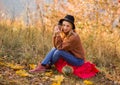 Female autumn portrait. cute blonde girl in a brown warm sweater, black felt hat, blue jeans sits on a plaid on the grass, holds a Royalty Free Stock Photo