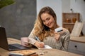 Female blogger in beige sweater. Smiling, sitting in kitchen at table, talking by smartphone, going write something in Royalty Free Stock Photo