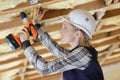 Female builder using cordless drill on wooden ceiling joist